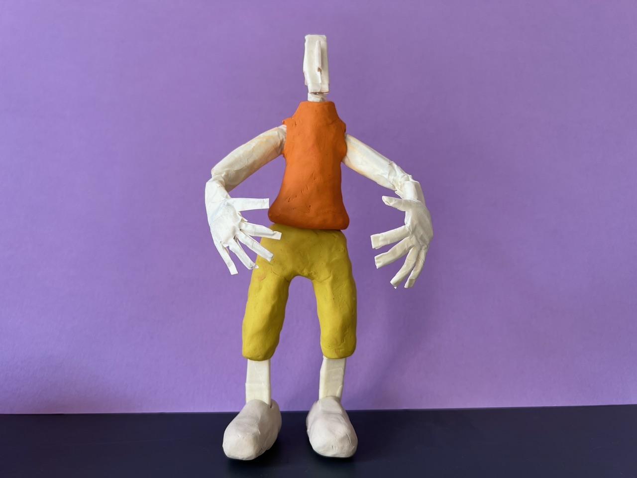Animated creature infront of a purple wall with their arms forward an orange plastecine tummy and yellow trousers