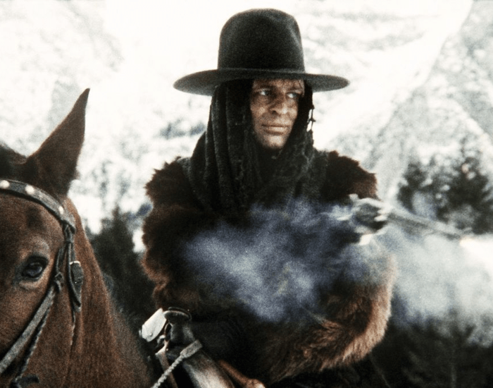 A sinister-looking man dressed in dark clothing fires a gun on horseback