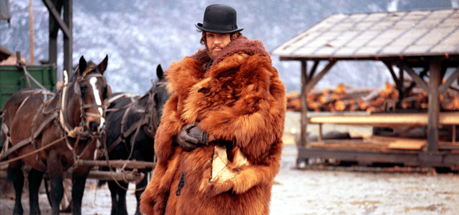 A man wears a big fur coat and a bowler-style hat and stands outside in a wet Old West town