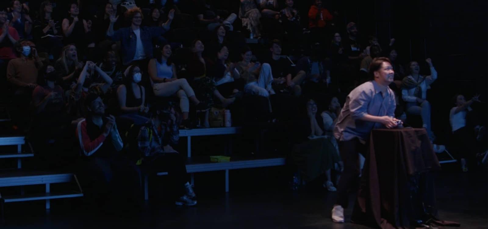 Promotional image for asses.masses showing an audience member playing the videogame in front of an audience