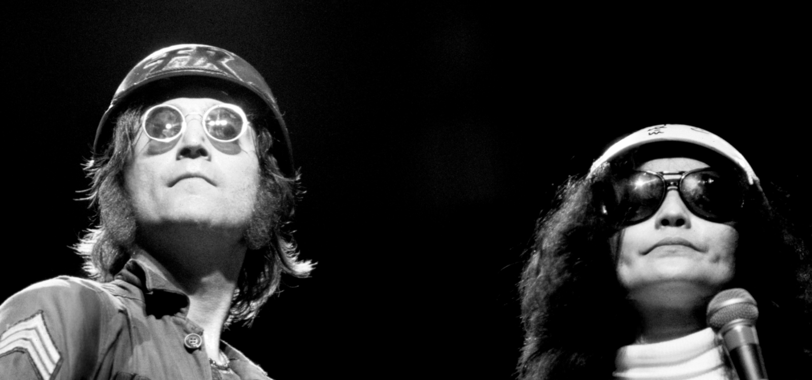Black-and-white photo of John Lennon and Yoko Ono on stage wearing helmets and dark sunglasses. The person on the left holds an electric guitar and looks upward, while the person on the right holds a microphone and faces forward. The background is dark, with stage lighting highlighting their faces and upper bodies.
