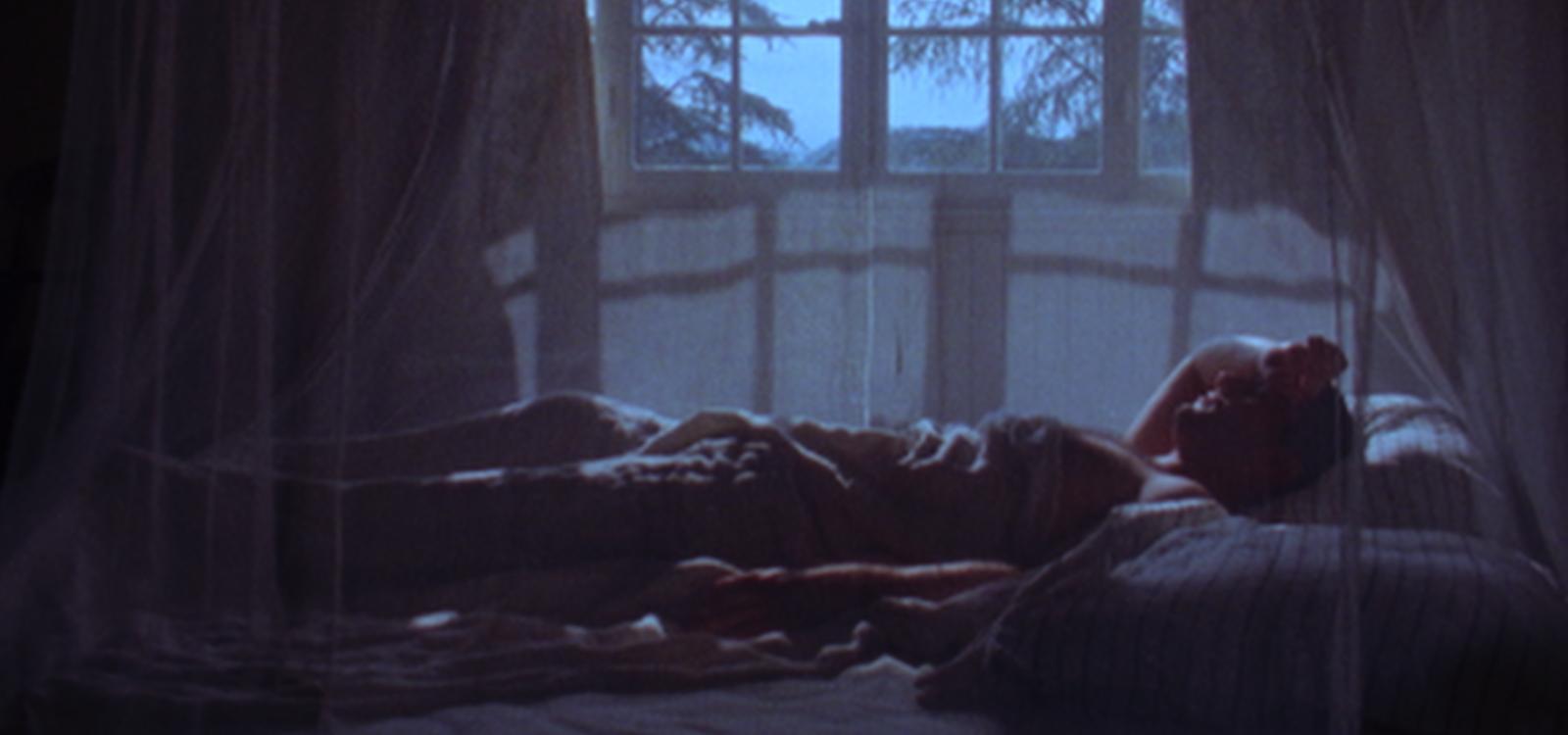 Chanthila Phaophanit, Beneath the Shell of Sound, 2025 In a dimly lit bedroom, a person lies on a bed beneath a translucent mosquito net canopy. Blue moonlight enters through a large-paned window behind them, casting soft shadows across the room. Silhouettes of trees are visible outside, while the figure rests with one arm bent behind their head, creating a quiet, contemplative atmosphere. The image has a dreamlike, cinematic quality, with deep blues and soft focus enhancing the sense of solitude and reflec