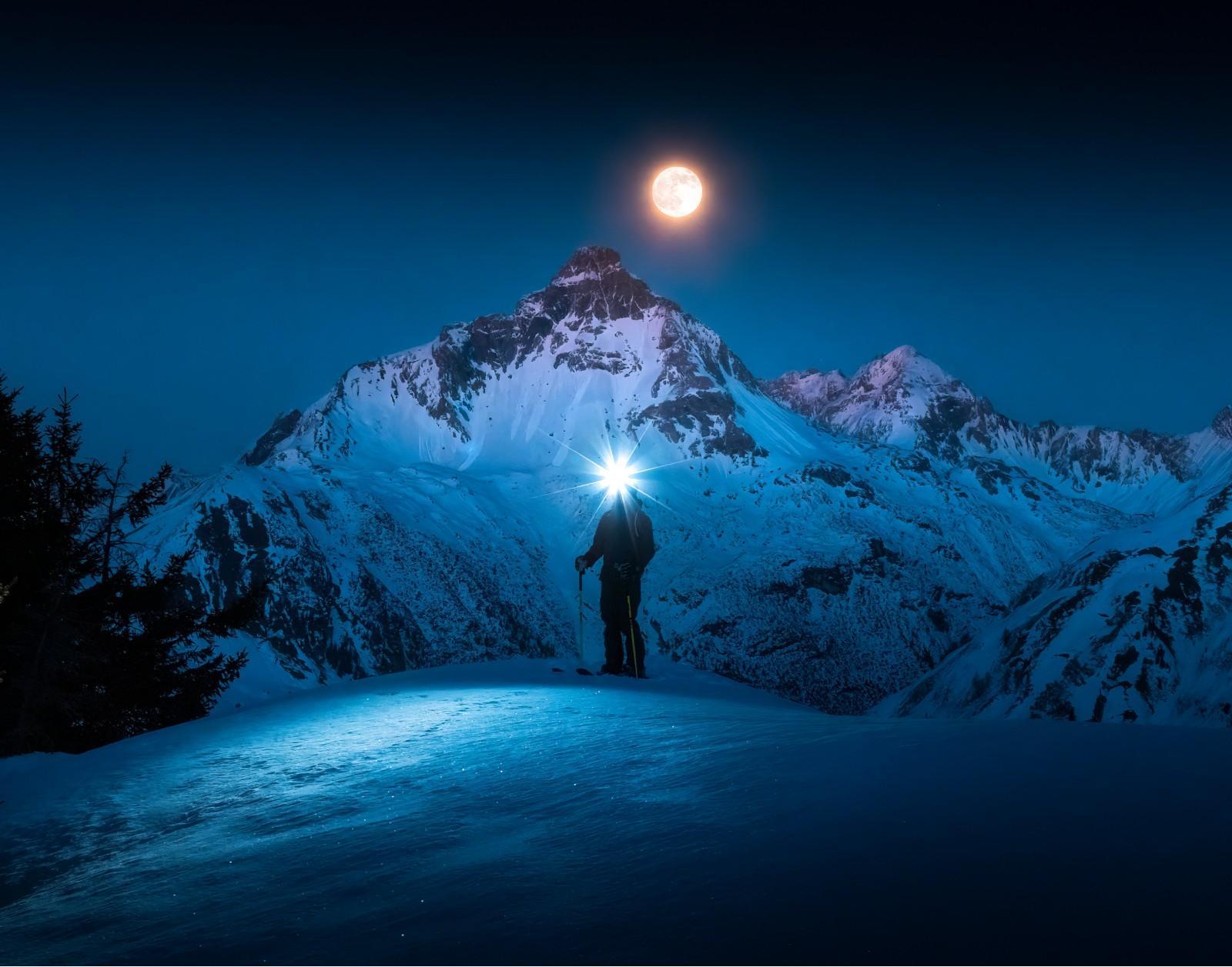 A person with a headlamp stands on a snowy mountain at night, silhouetted against a distant peak under a full moon.