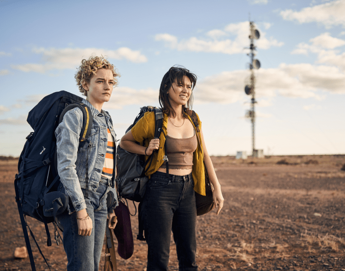 Two young women wearing backpacks stand in a dusty Outback landscape