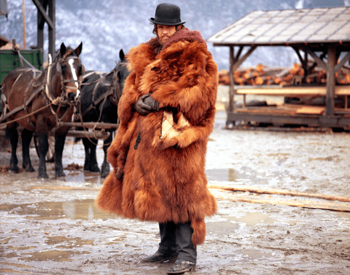 A man wears a big fur coat and a bowler-style hat and stands outside in a wet Old West town