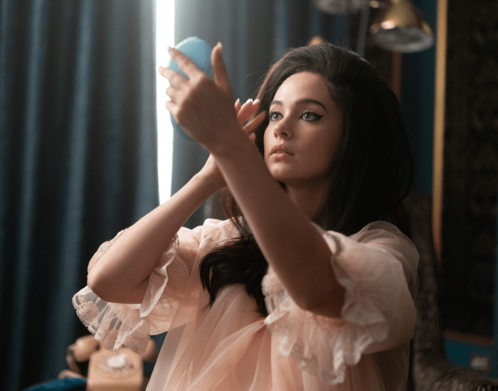 A young woman wearing a nightgown with styled hair and make-up looks at her reflection in a compact mirror