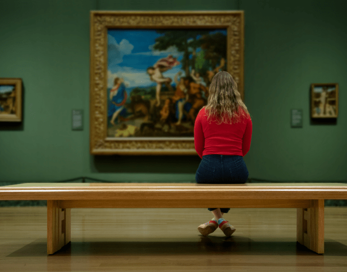 A woman sits on a bench in front of a large painting in a gallery
