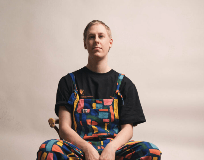 Joe Egan sat on a wooden chair in multi coloured dungarees 