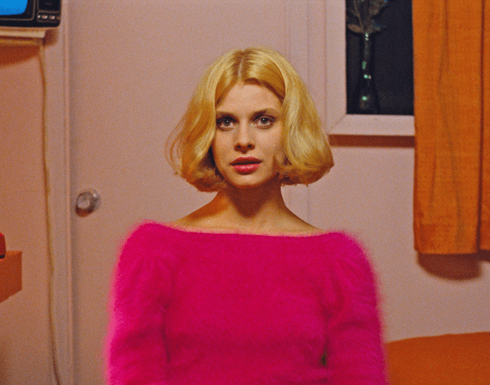 A young blonde woman sits dressed in vibrant fuschia looking towards the camera