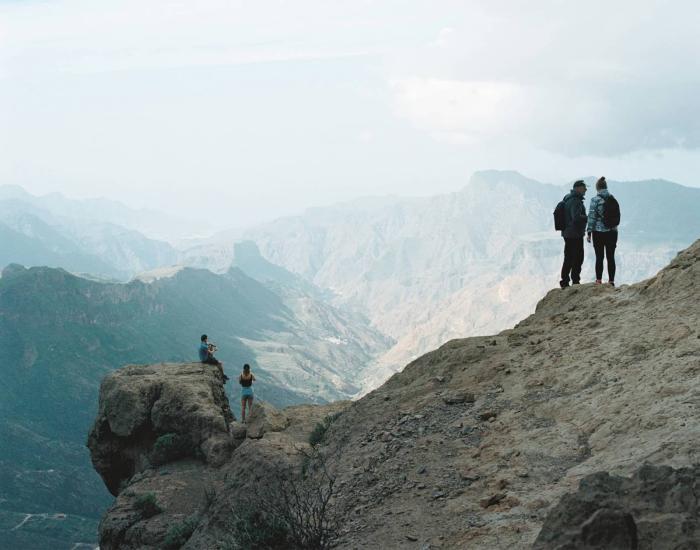 Two people stand at the top of a mountain looking out to a nearby peak, two people are closeby slightly lower down  sat on a mound