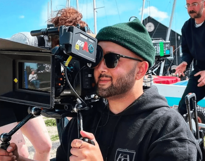 Jordan Heath holding a camera looking into the lens whilst wearing sunglasses and a green wooley hat