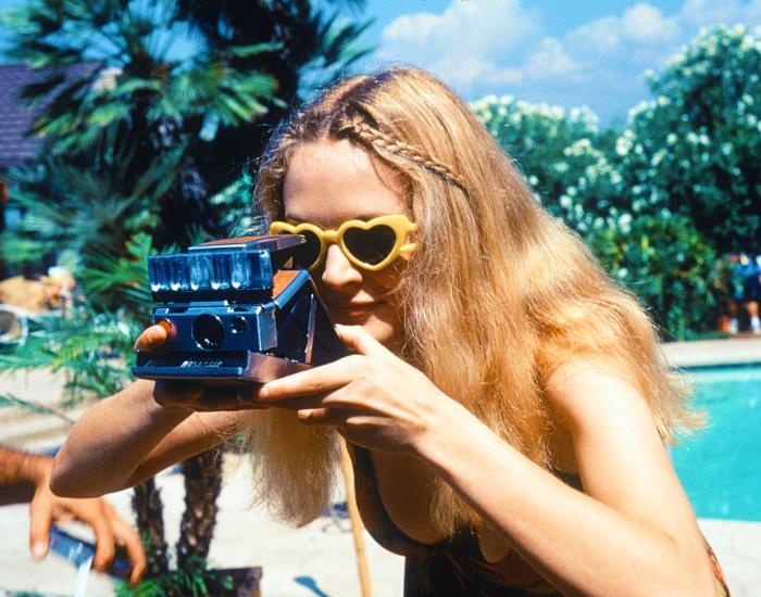 A young woman wearing sunglasses beside a pool takes a Polaroid photograph